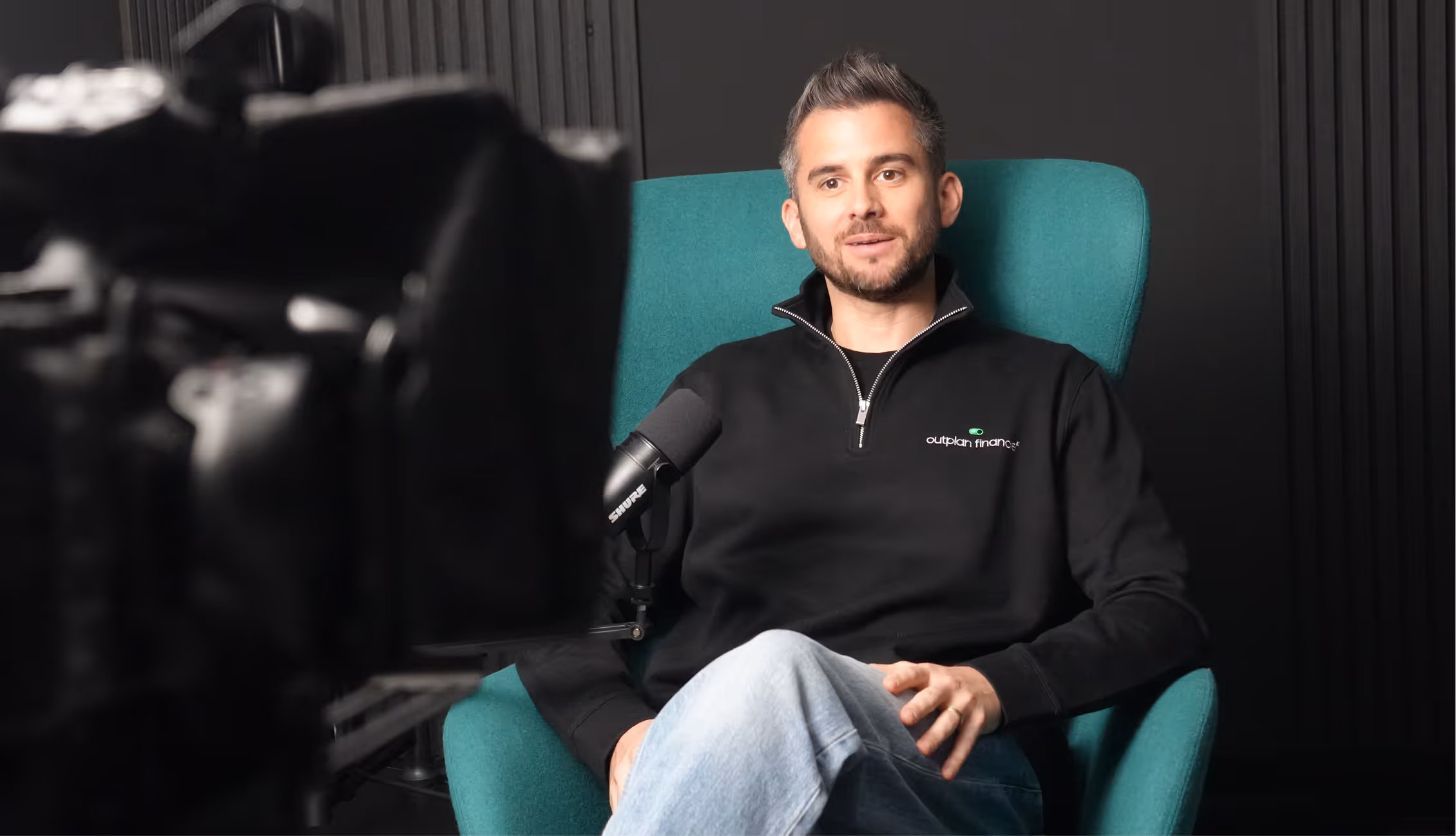 Man with short hair and beard sitting in a teal armchair, wearing a black Outplan Finance sweatshirt, speaking into a microphone during a video recording.