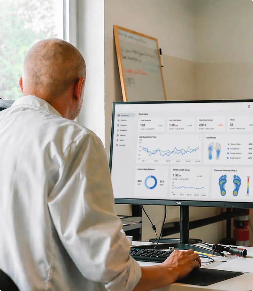 Man in white shirt working on a computer displaying gait analysis charts and foot pressure heatmaps.