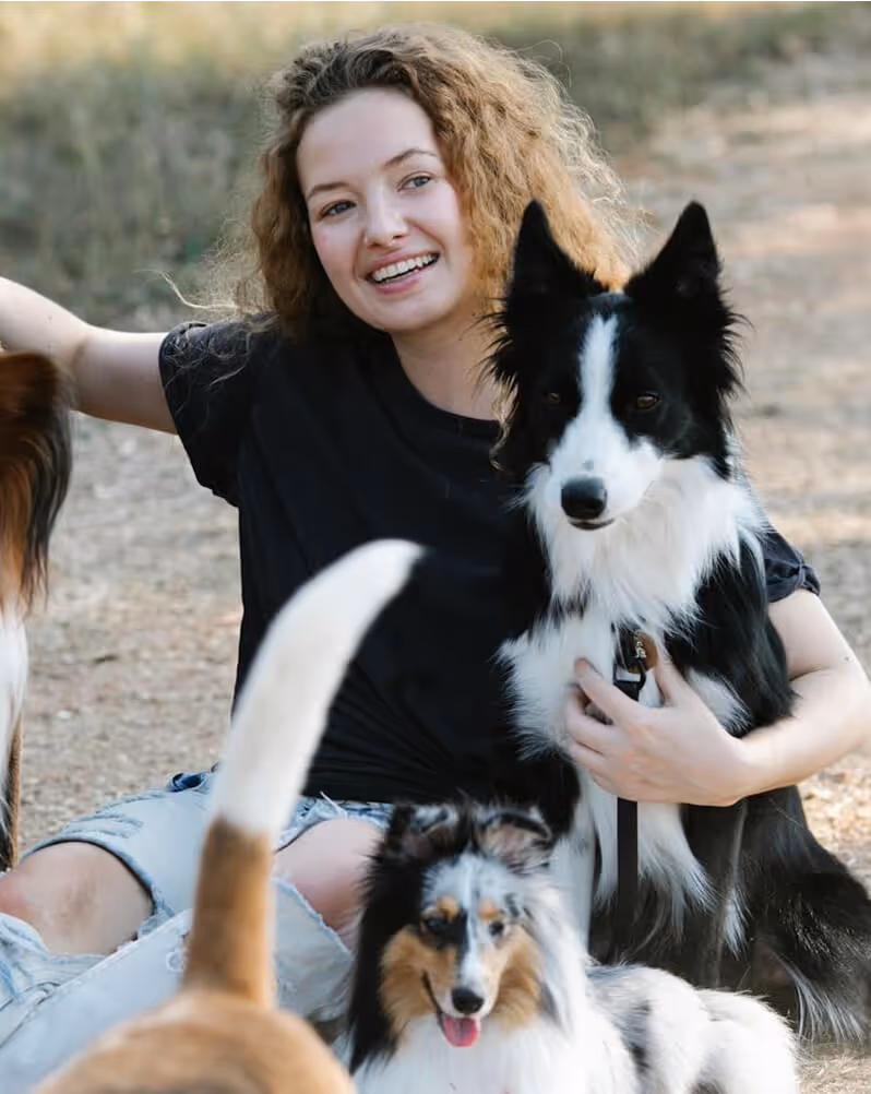 Smiling woman sitting outdoors with three dogs, holding a black and white dog close.