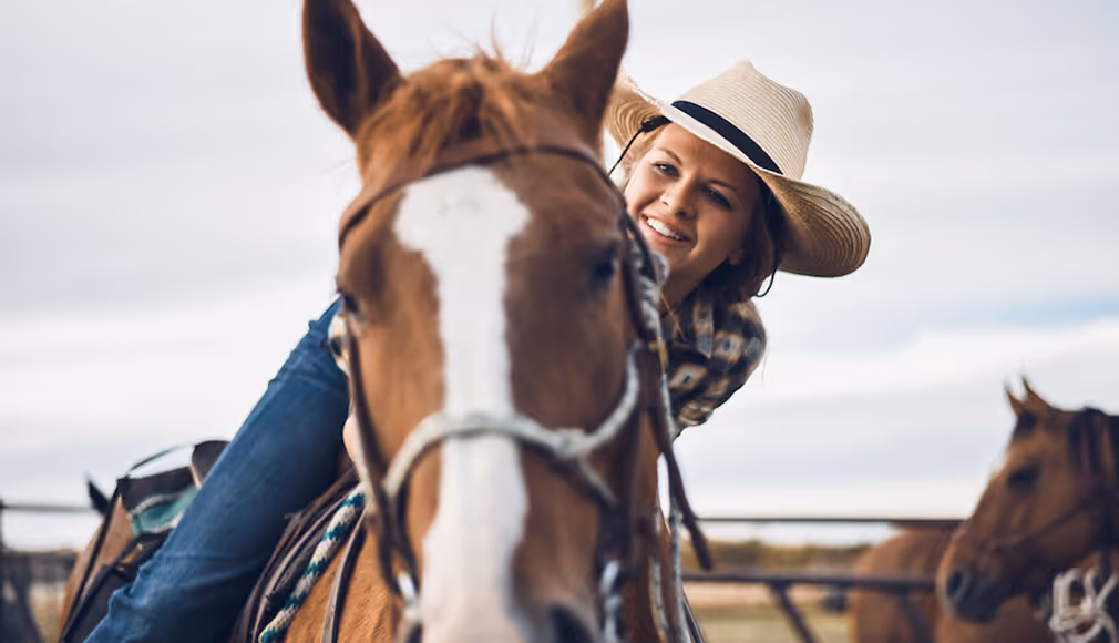 Smiling woman in a hat riding a brown horse outdoors with other horses in the background.