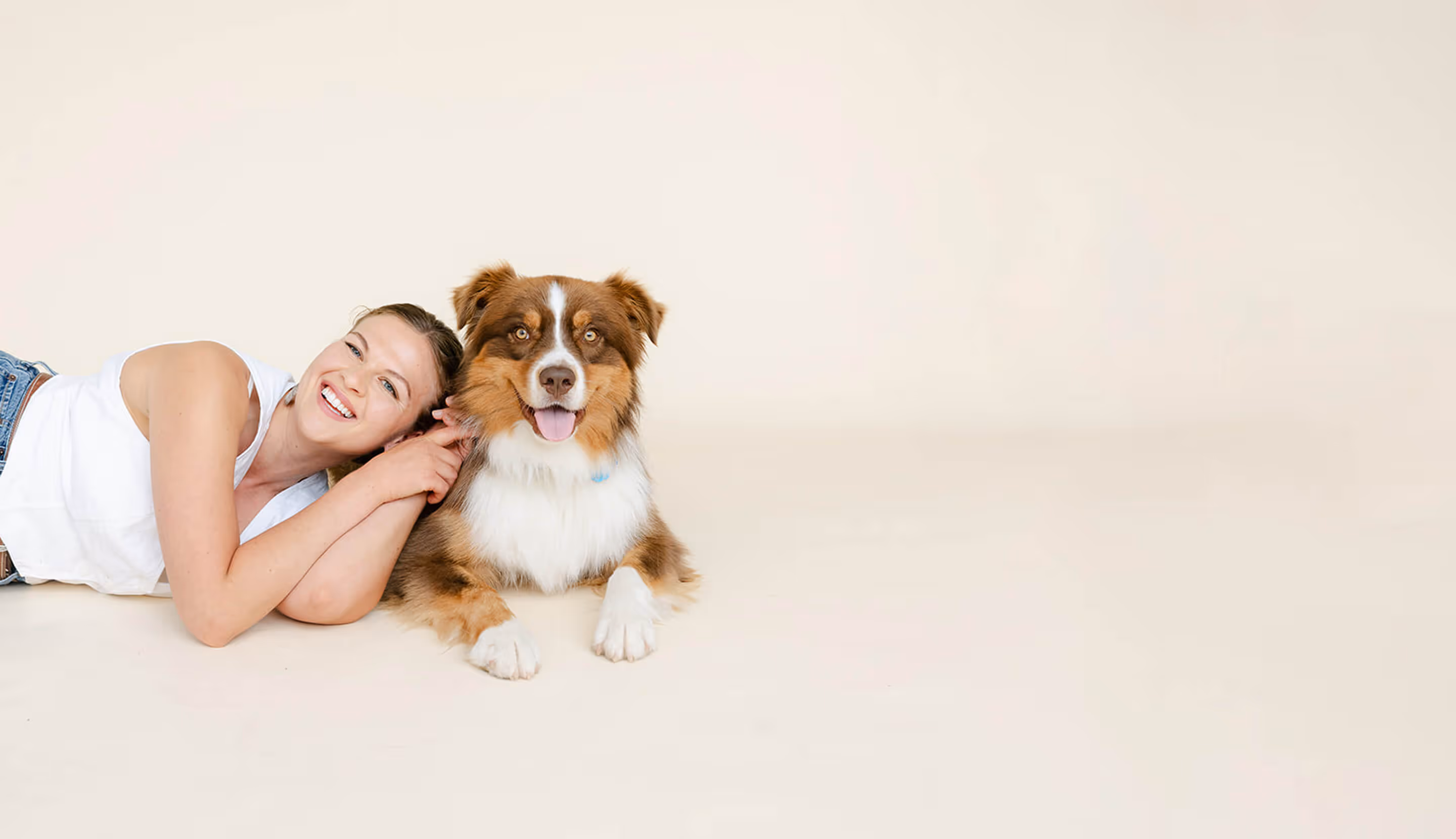 Smiling woman lying on floor next to a happy brown and white dog against beige background.