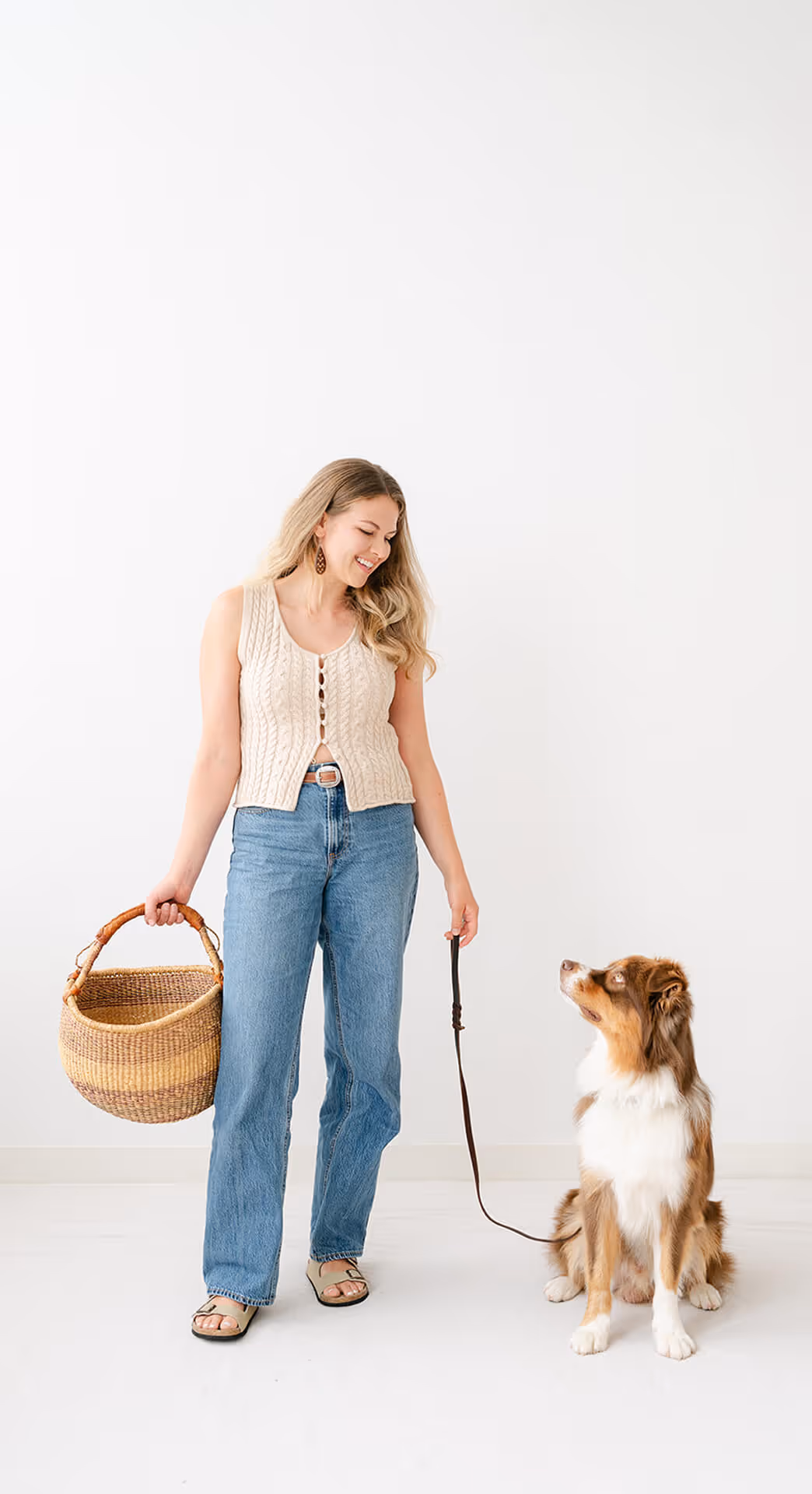Smiling woman holding a basket and leash, standing next to a sitting brown and white dog.