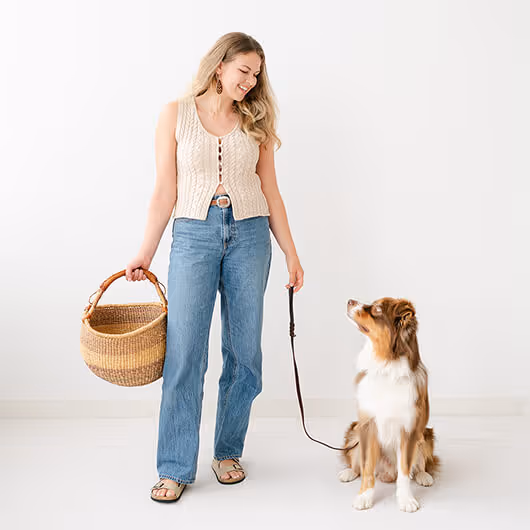 Woman smiling holding basket and leash while looking at a seated Australian Shepherd dog.