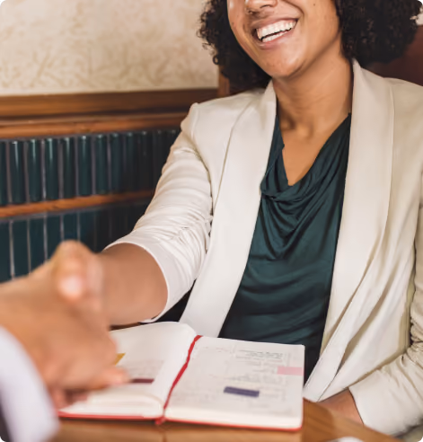 Smiling person in a beige blazer shaking hands across a table with an open planner.