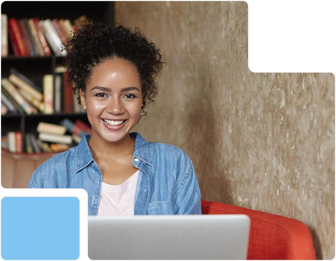 Smiling young woman with curly hair in a denim shirt sitting on an orange couch with a laptop in front of her and a bookshelf in the background.