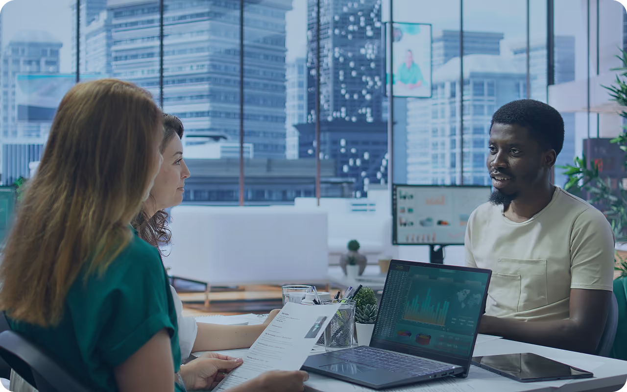 Three people having a discussion in a modern office with a cityscape view, with a laptop displaying graphs and charts on the table.