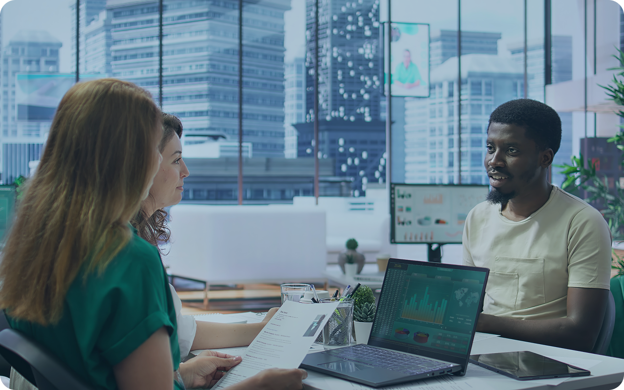 Three people having a discussion in a modern office with a cityscape view, with a laptop displaying graphs and charts on the table.