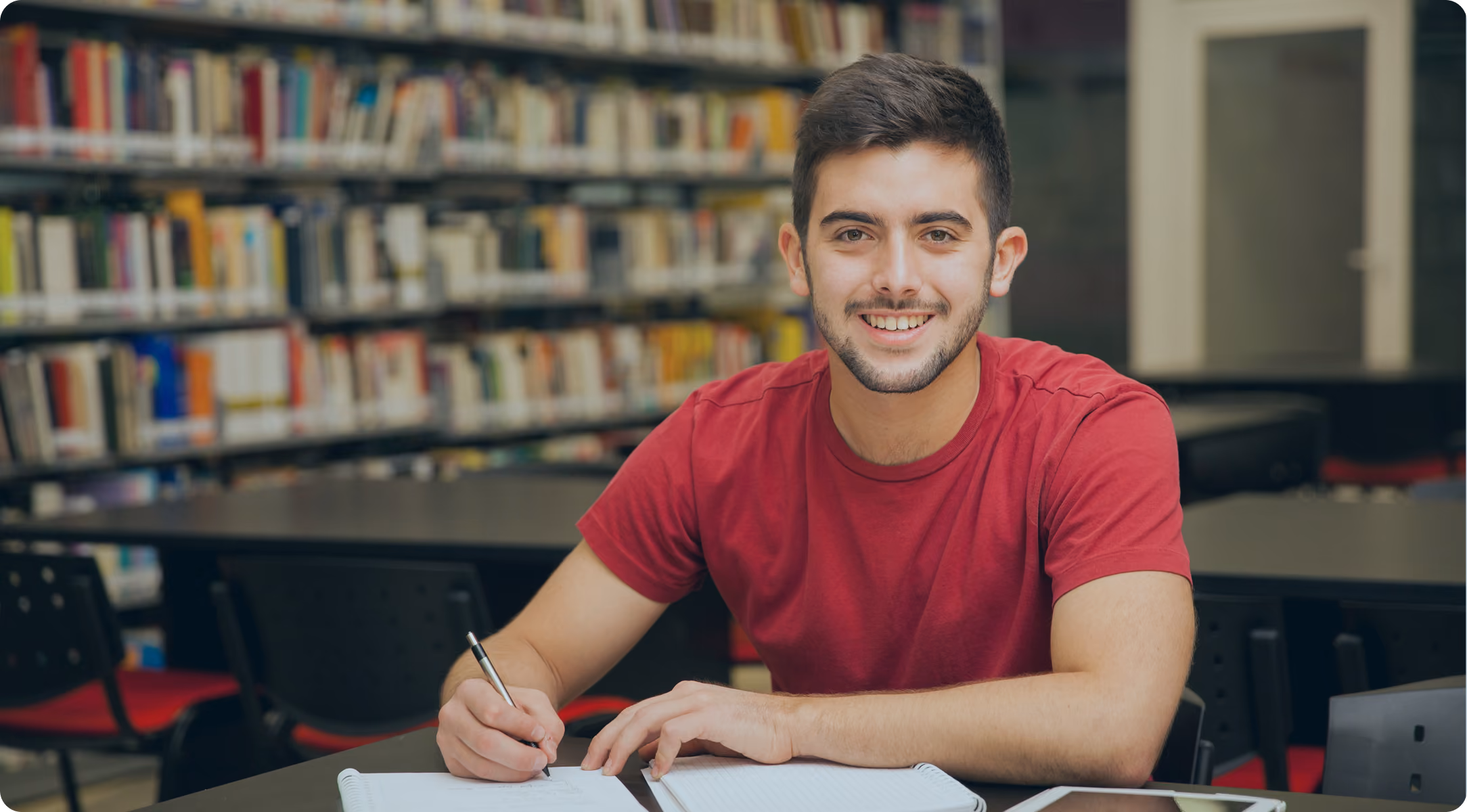 Smiling young man in a red shirt writing in a notebook inside a library.
