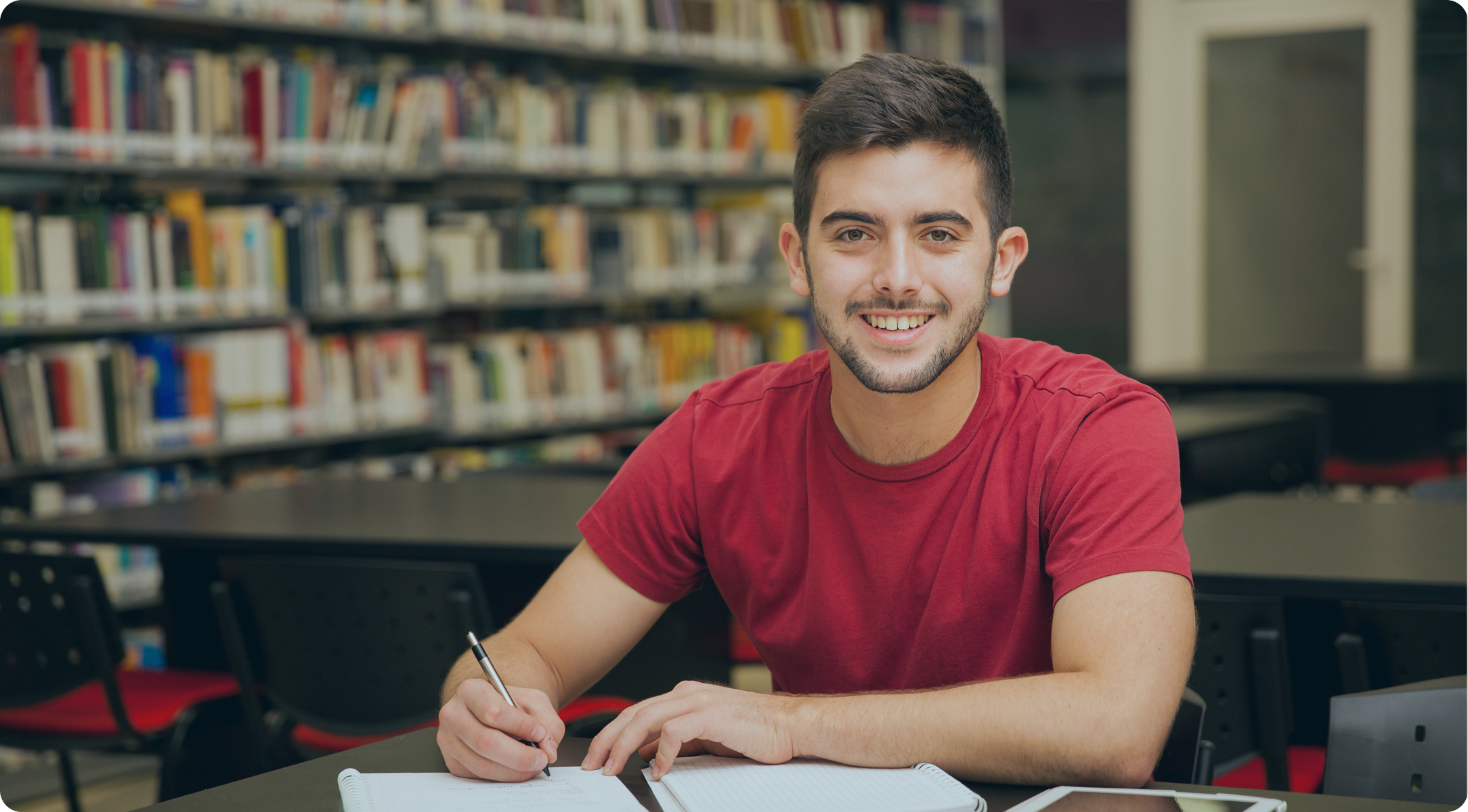 Smiling young man in a red shirt writing in a notebook inside a library.
