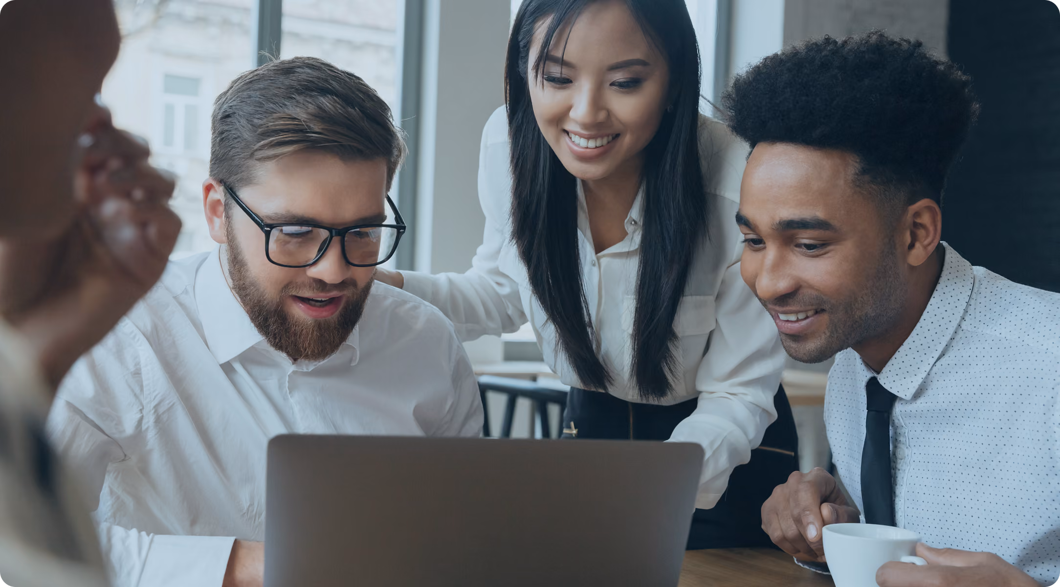 Three diverse colleagues smiling and collaborating while looking at a laptop screen in an office.