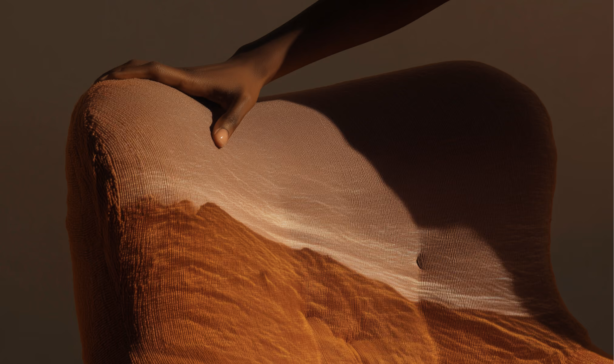 Hand resting on a fabric chairback lit by warm light creating mountain-like shadows.