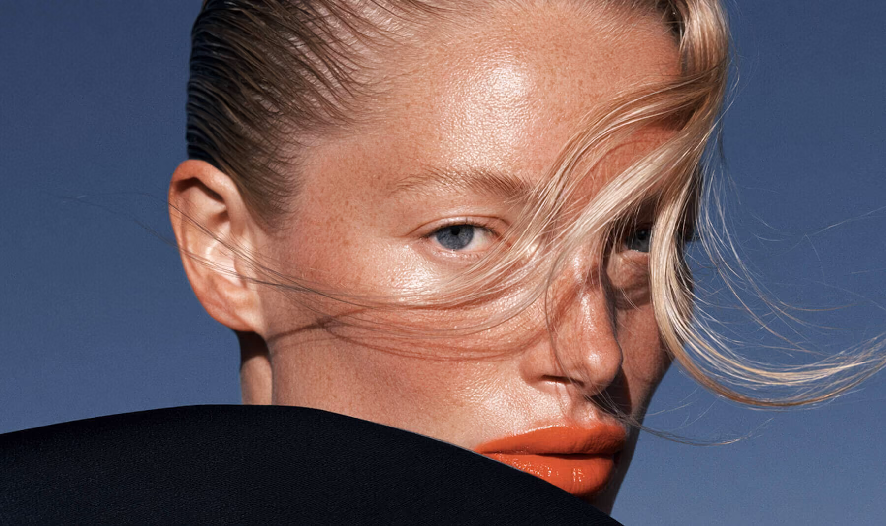 Close-up of a woman with slicked-back hair, light eyes, orange lipstick, and strands of hair across her face.