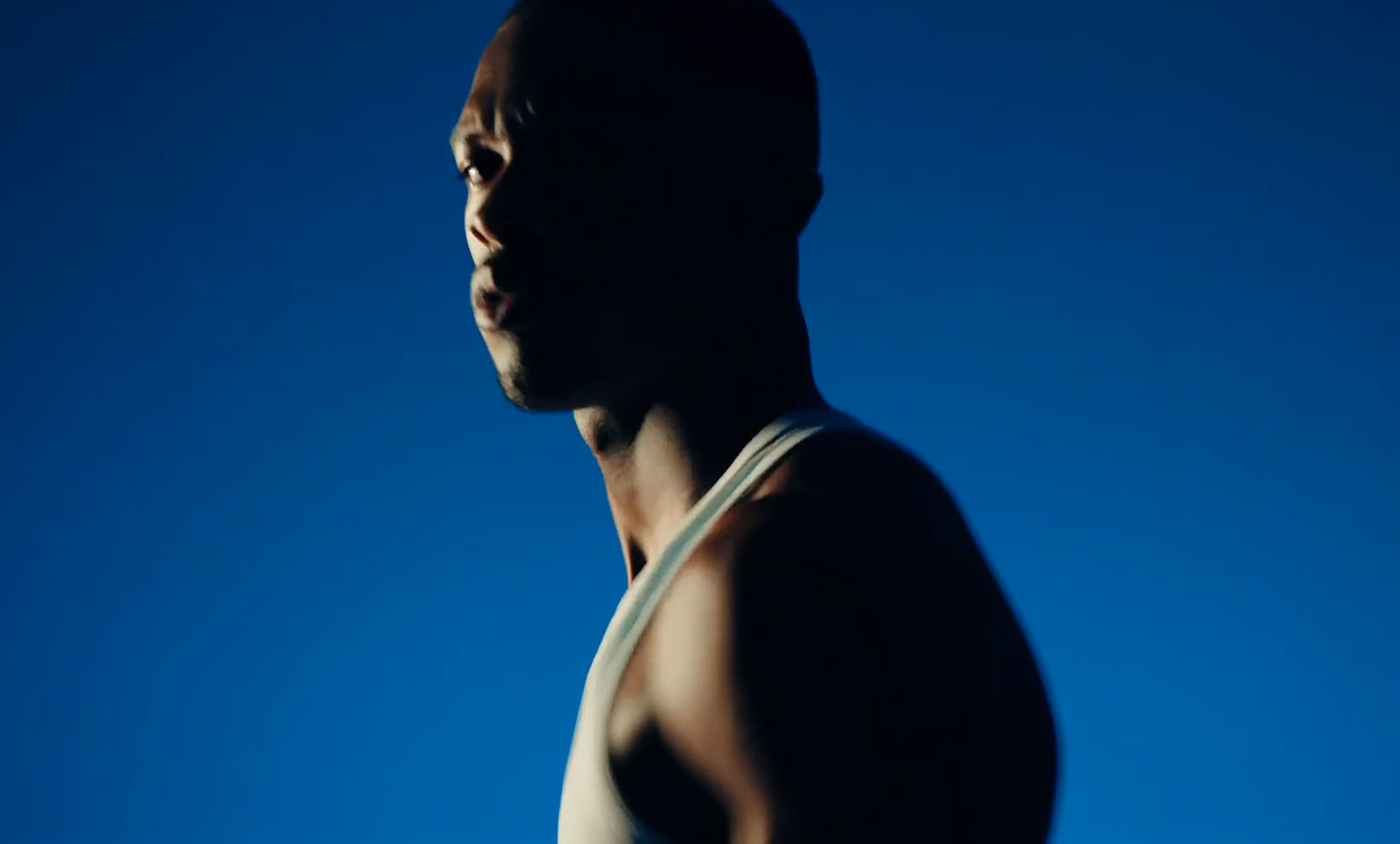 Man in white tank top looking to the side with a dark blue background.