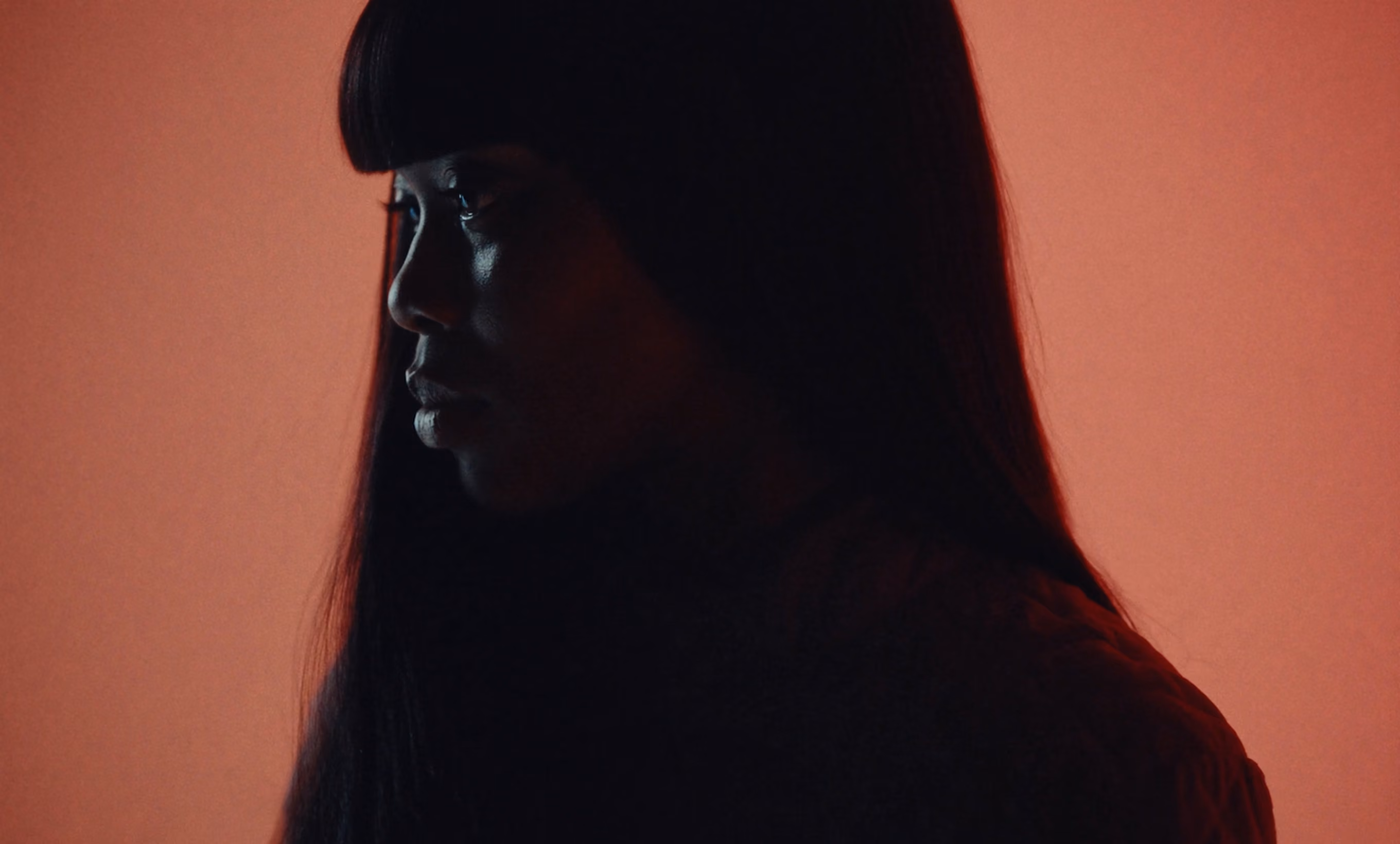 Side profile of a woman with long hair against a reddish-orange background in low light.