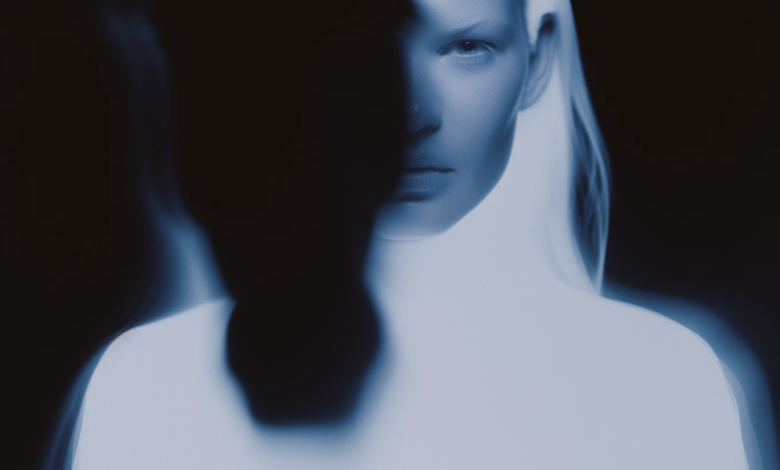 Blurred dark silhouette facing a sharp blue-tinged woman’s face in a dark background.