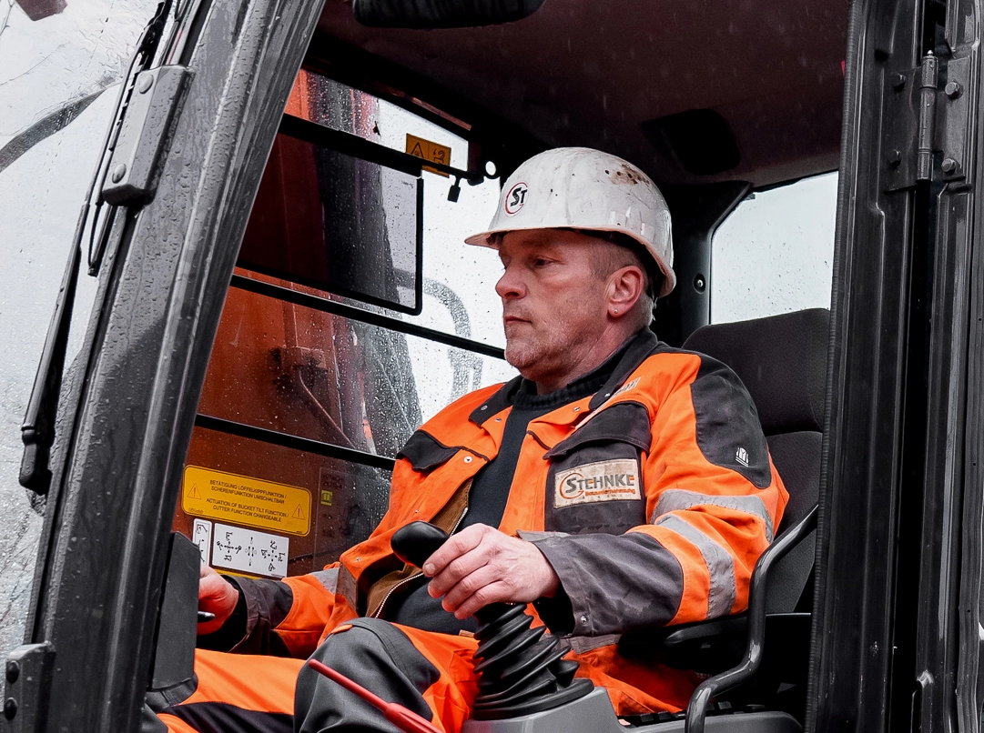 Bauarbeiter mit orangefarbener Warnschutzjacke und weißem Helm sitzt in der Kabine eines Fahrzeugs und bedient die Steuerhebel.