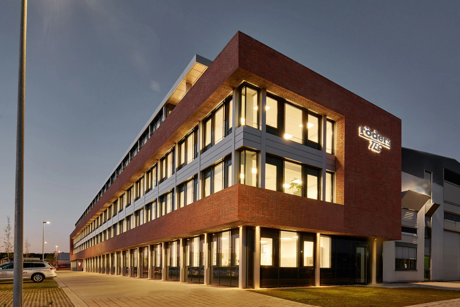 Modernes Bürogebäude mit roter Backsteinfassade und beleuchteten Fenstern bei Dämmerung mit dem Schriftzug röders TEC.