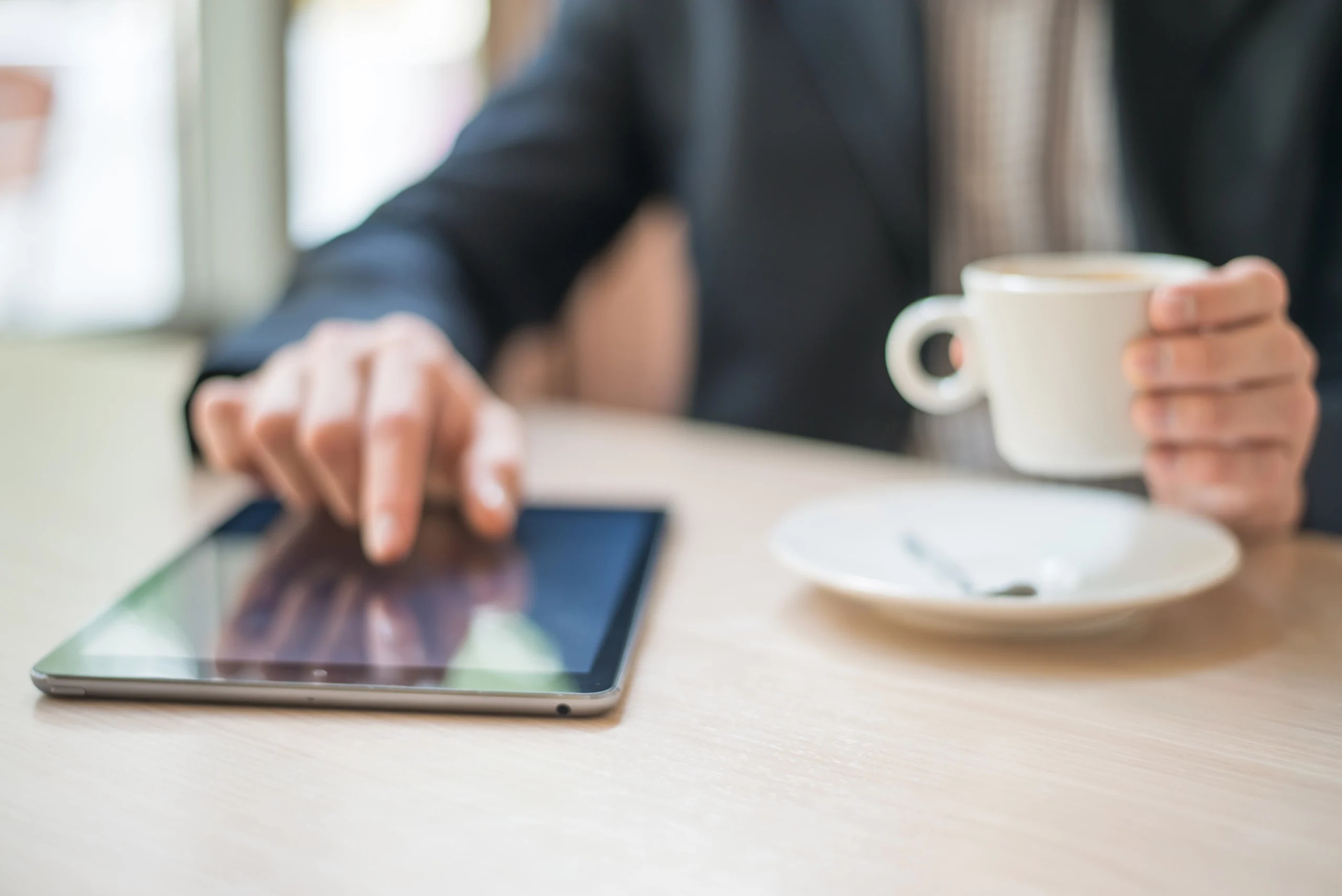Person hält eine weiße Tasse Kaffee und bedient mit der anderen Hand ein Tablet auf einem Tisch.