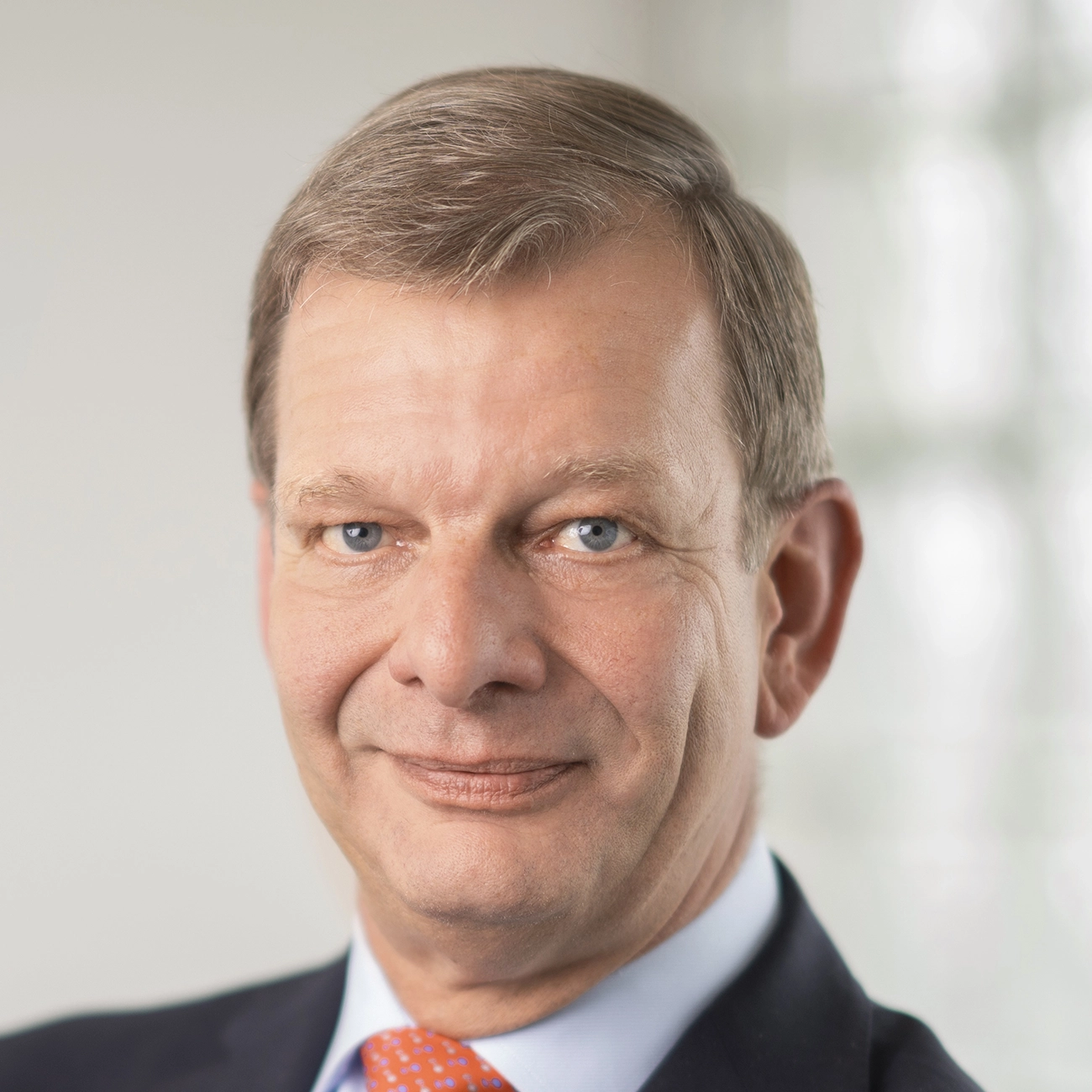 Porträt eines lächelnden älteren Mannes mit blauen Augen, braunem Haar, einem dunklen Anzug, hellblauem Hemd und orangefarbener Krawatte.