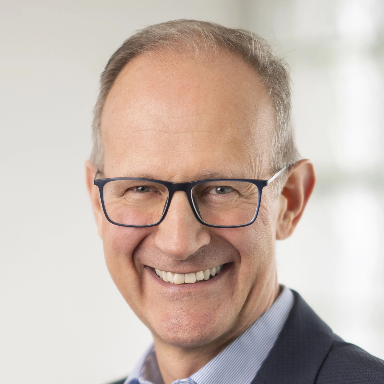 Lächelnder älterer Mann mit Brille und Anzug vor hellem Hintergrund.