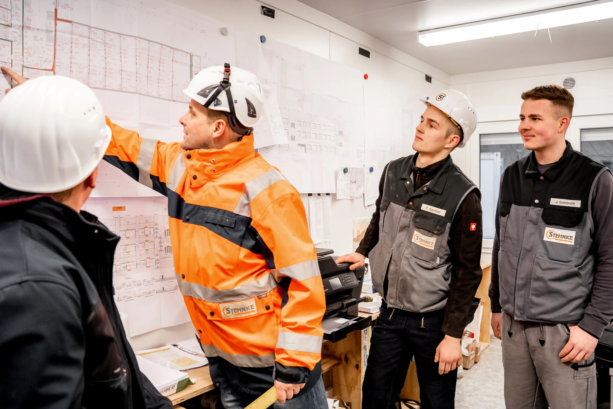 Vier Bauarbeiter mit Schutzhelmen besprechen Pläne an einer Wand in einem Büro.