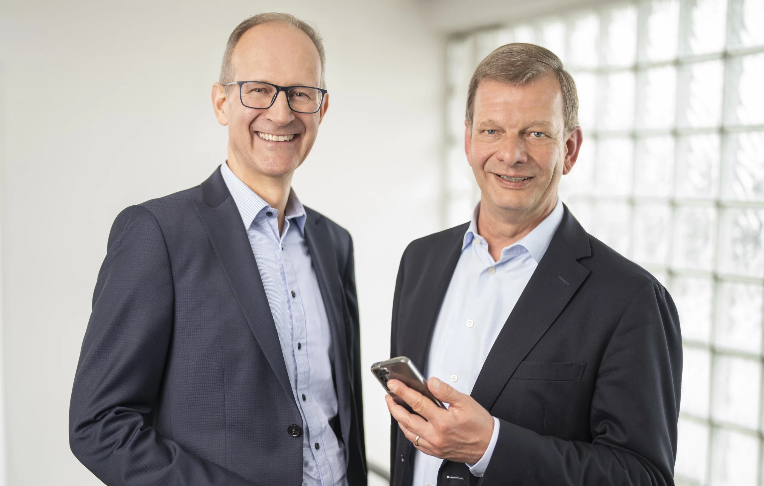 Zwei lächelnde Männer in Anzügen, einer hält ein Smartphone, vor hellem Hintergrund mit Glasfenster.