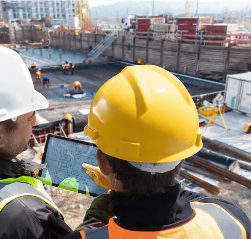 Zwei Bauarbeiter mit Schutzhelmen betrachten einen Bauplan auf einem Tablet an einer Baustelle.