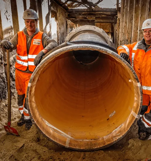 Zwei Bauarbeiter in orangen Warnwesten neben einer großen, liegenden Rohrleitung auf einer Baustelle.