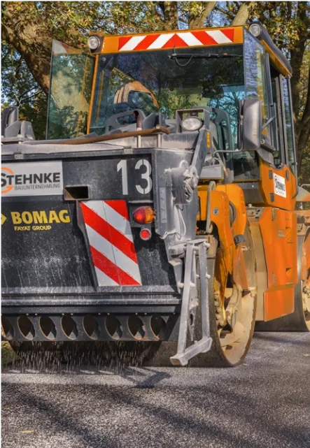 Nahaufnahme eines orangefarbenen Bomag Straßenwalzenfahrzeugs, das Asphalt auf einer Straße glättet.