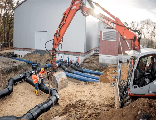 Baustelle mit orangefarbenem Bagger und Arbeitern, die große Rohrleitungen verlegen.