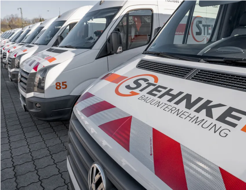 Reihe von weißen Volkswagen Transportern mit Stehnke Bauunternehmung Schriftzug und rotem Warnmuster vorne.