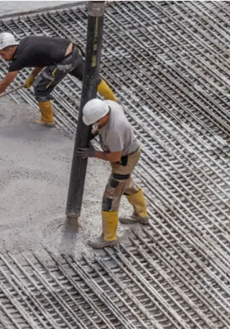 Baustellenarbeiter gießen Beton über ein Stahlbewehrungsnetz mit gelben Gummistiefeln und Schutzhelmen.