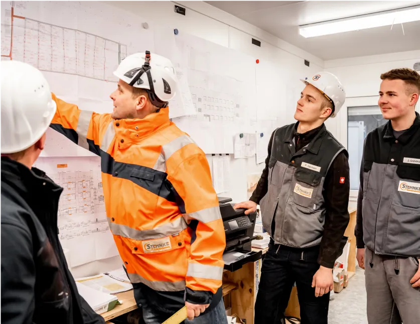 Vier Bauarbeiter mit Schutzhelmen besprechen Pläne an einer Wand mit Bauzeichnungen in einem Büro.