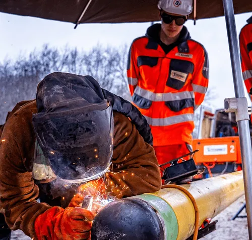 Arbeiter in Schutzkleidung schweißt draußen eine große Pipeline, im Hintergrund steht eine zweite Person in Sicherheitsanzug.