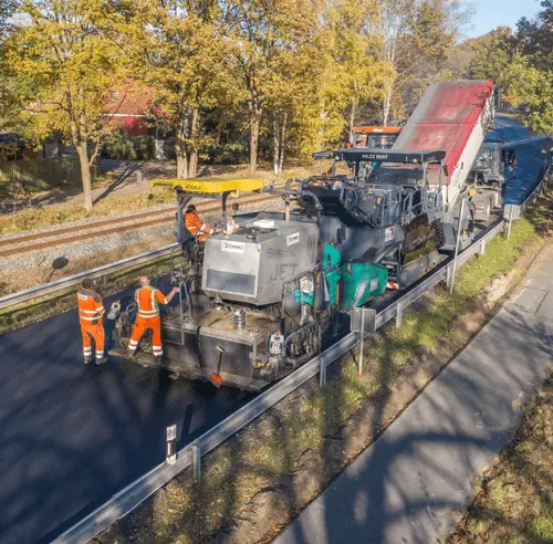 Straßenarbeiter in orangen Warnwesten bedienen eine Straßenfertigmaschine, die Asphalt auf einer Straße aufträgt.