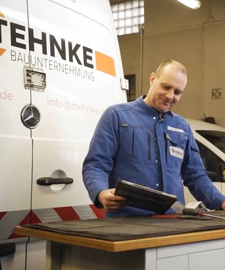 Ein Mann in blauer Arbeitsjacke steht vor einem Fahrzeug mit Stehnke-Bauunternehmenslogo und hält ein Tablet.