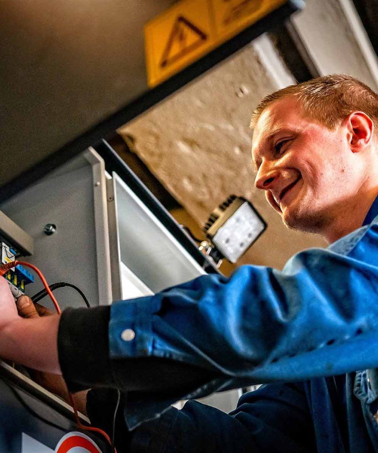 Mann im blauen Arbeitsanzug arbeitet lächelnd an einem elektrischen Schaltschrank.