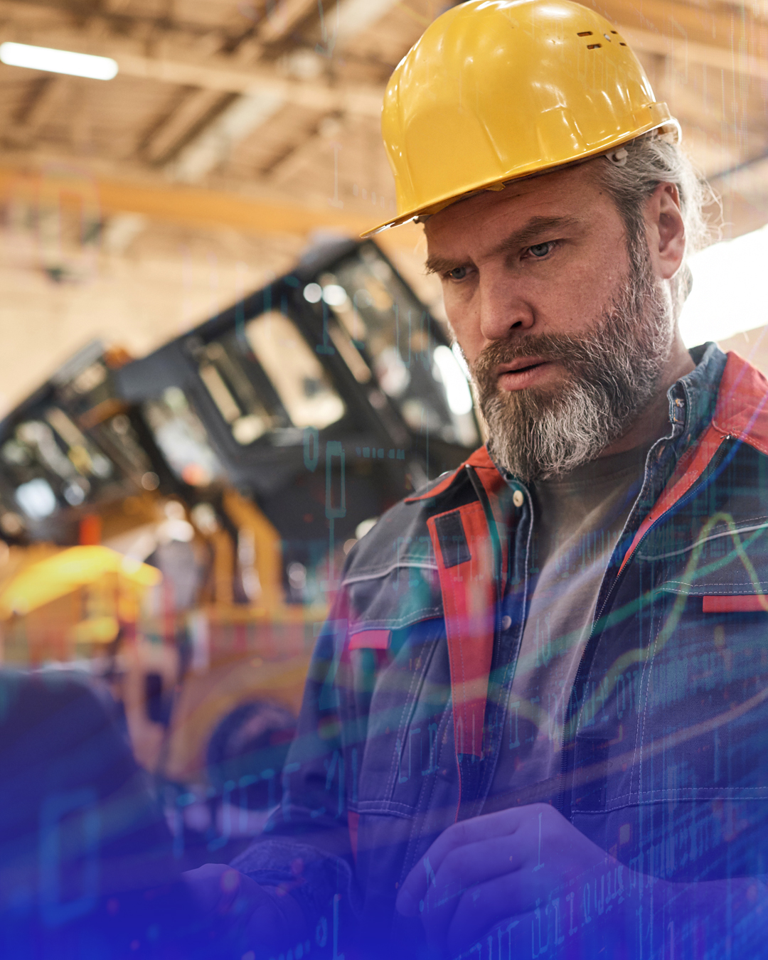 Bearded man in a yellow hardhat and work jacket looking down with a serious expression in an industrial setting.