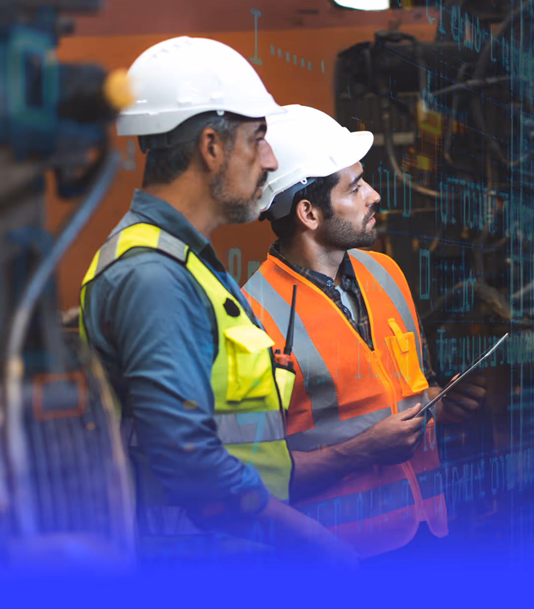 Two industrial workers wearing white hard hats and safety vests are focused on inspecting equipment.