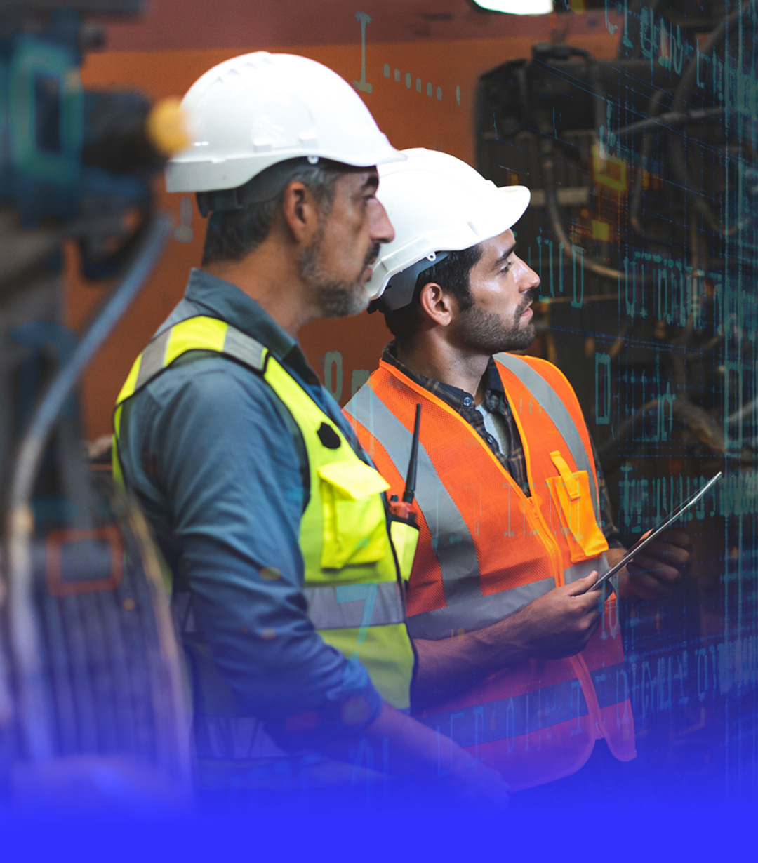 Two industrial workers wearing white hard hats and safety vests are focused on inspecting equipment.