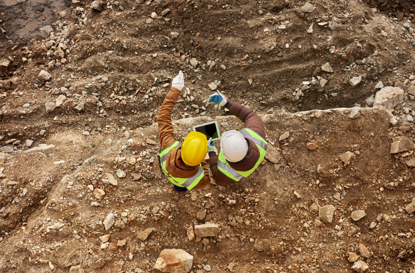 Two construction workers wearing helmets and reflective vests looking at a tablet on a rocky dirt site.