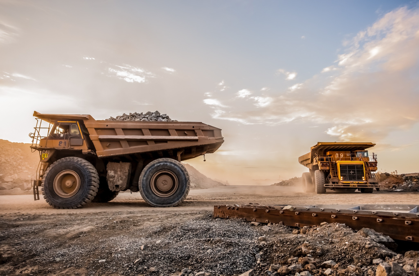 Two large yellow mining dump trucks on a dusty road in a quarry during sunset.
