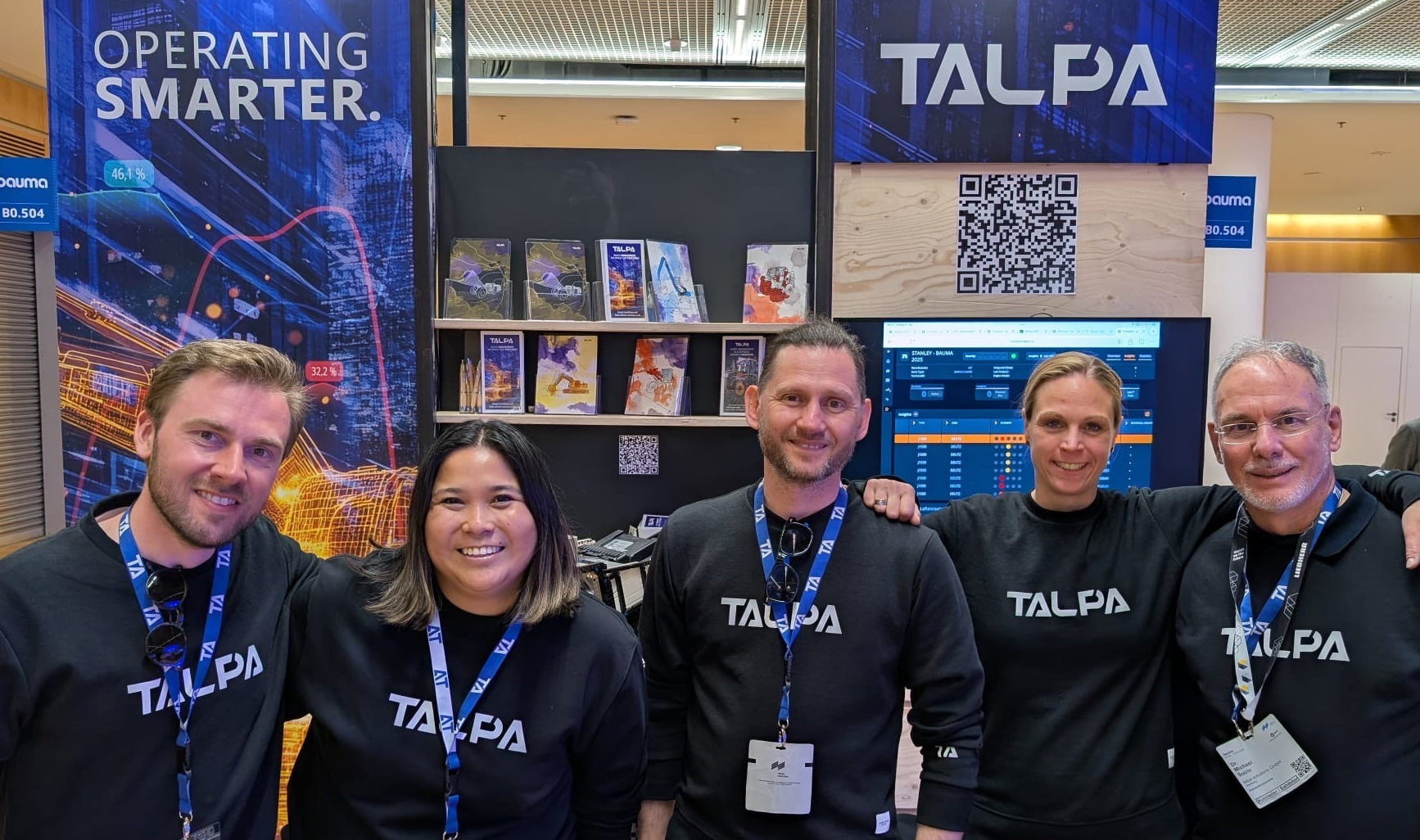 Five smiling team members wearing black TALPA shirts posing in front of a TALPA booth with posters and a screen.