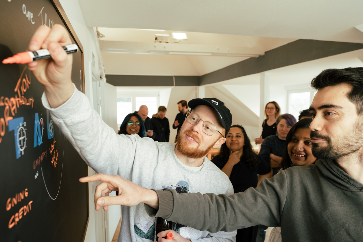 A TALPA team member in a gray sweatshirt and black cap writes on a blackboard with a red marker while another team member points and a group of people watch in a bright office room.
