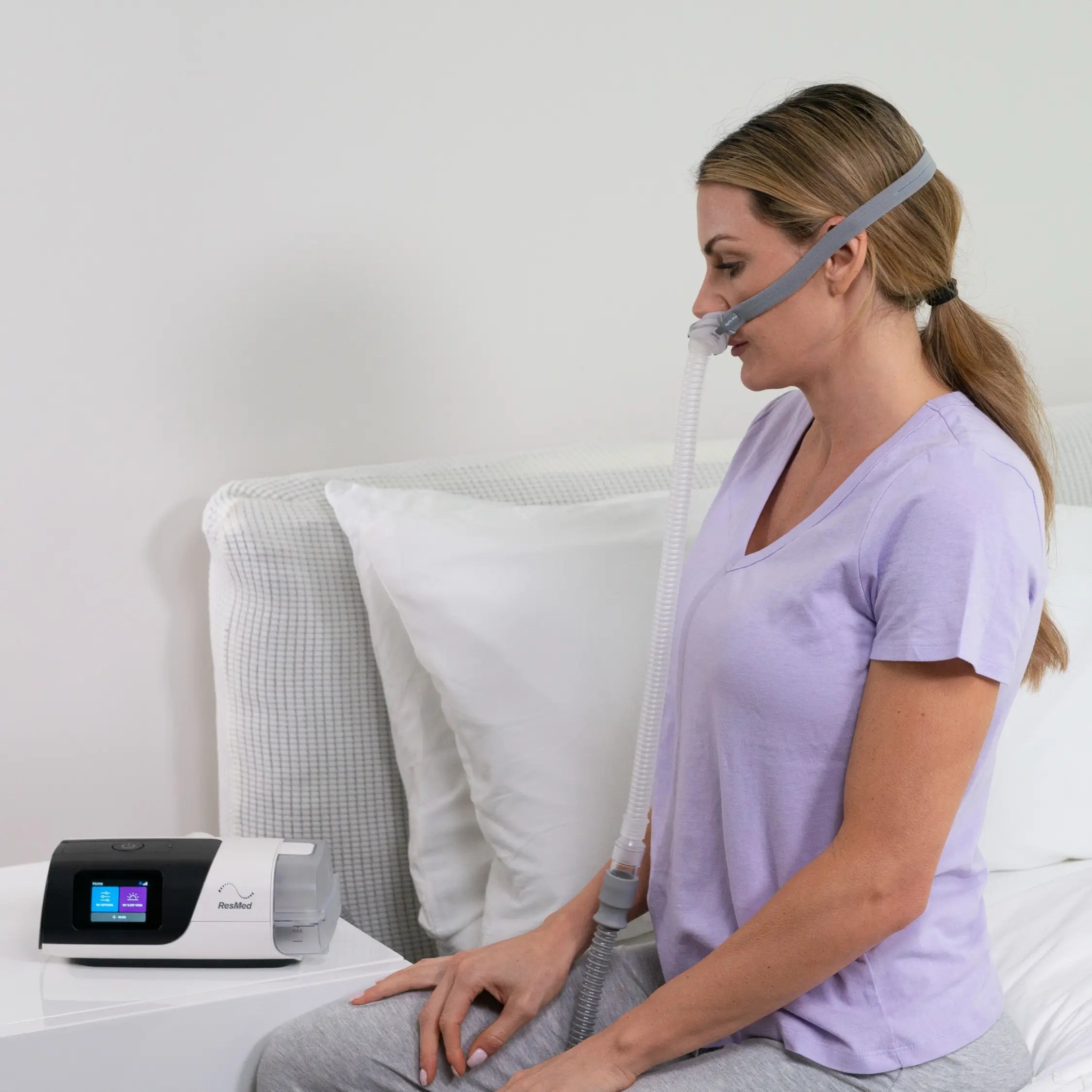 Woman sitting on a bed wearing a nasal mask connected to the ResMed AirSense 11