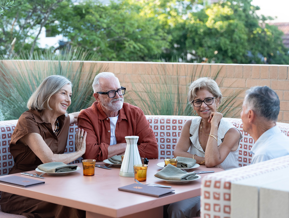 Four older adults seated outdoors around a table engaged in friendly conversation at a restaurant.