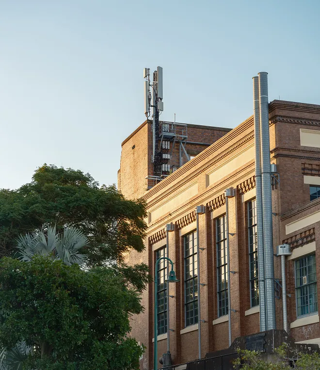 Brick building with tall windows and a cellular tower on the roof, beside green leafy trees.