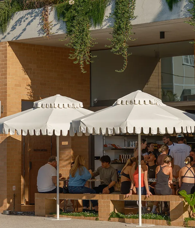 People sitting and socializing under white umbrellas outside a brick building with hanging plants above.