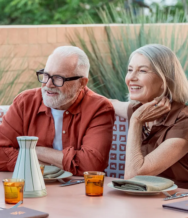 Smiling older couple sitting at a table outdoors with glasses of amber drink and tableware.