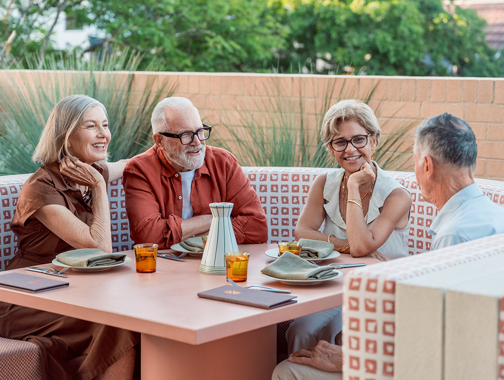 Four older adults seated outdoors around a table engaged in friendly conversation at a restaurant.